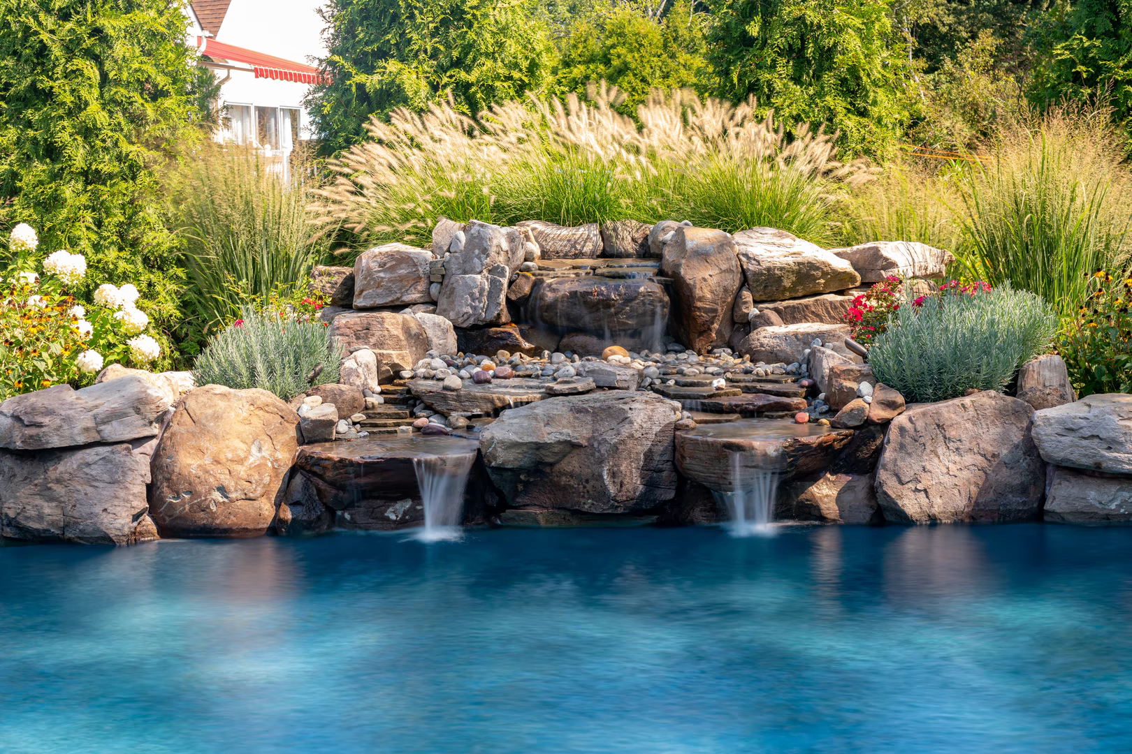 Natural stone boulder waterfall cascading into a pool surrounded by lush garden plants