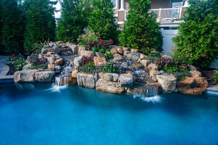Natural stone boulder waterfalls cascading into a swimming pool surrounded by lush landscaping