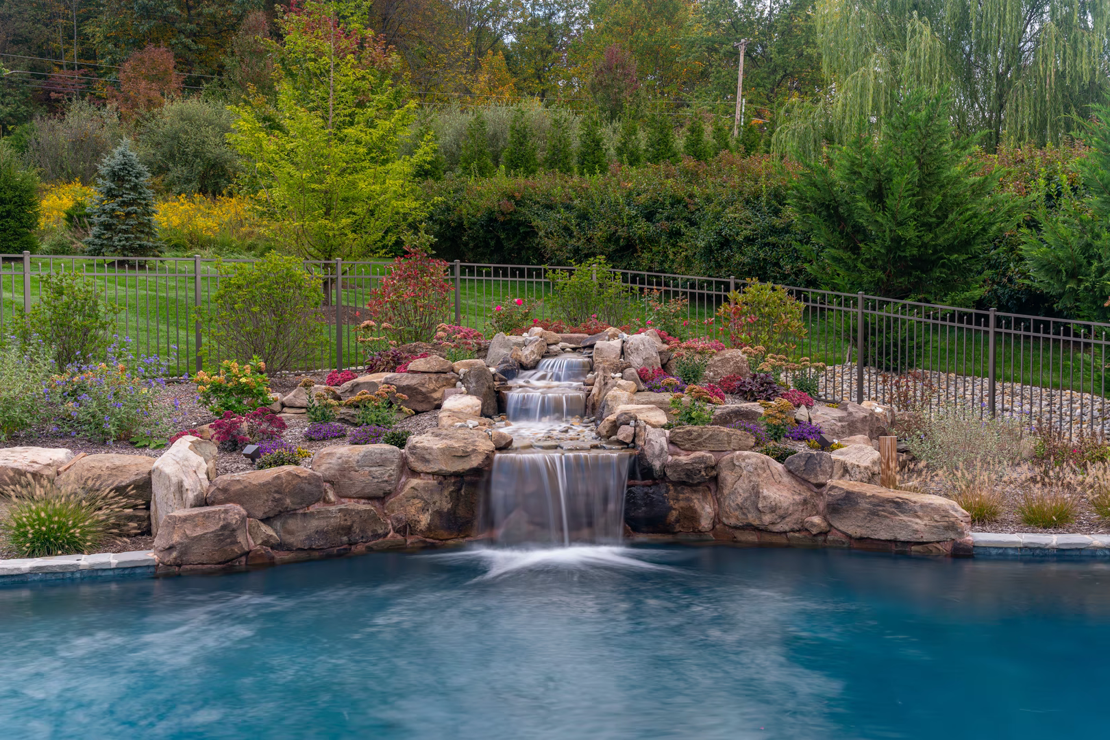 Cascading three-tier natural stone boulder waterfall flowing into a pool with colorful garden plants