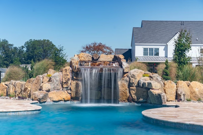 Kamui B - Phoenix, MD - A large rock waterfall in the middle of a swimming pool surrounded by lush greenery and a house in the background