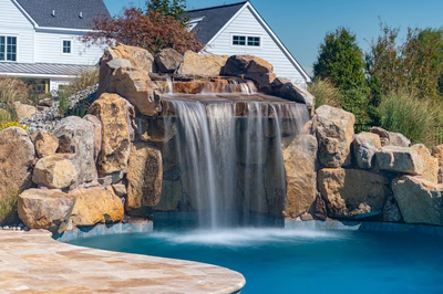Kamui C - Phoenix, MD - A large rock waterfall in a swimming pool surrounded by lush greenery and a house in the background