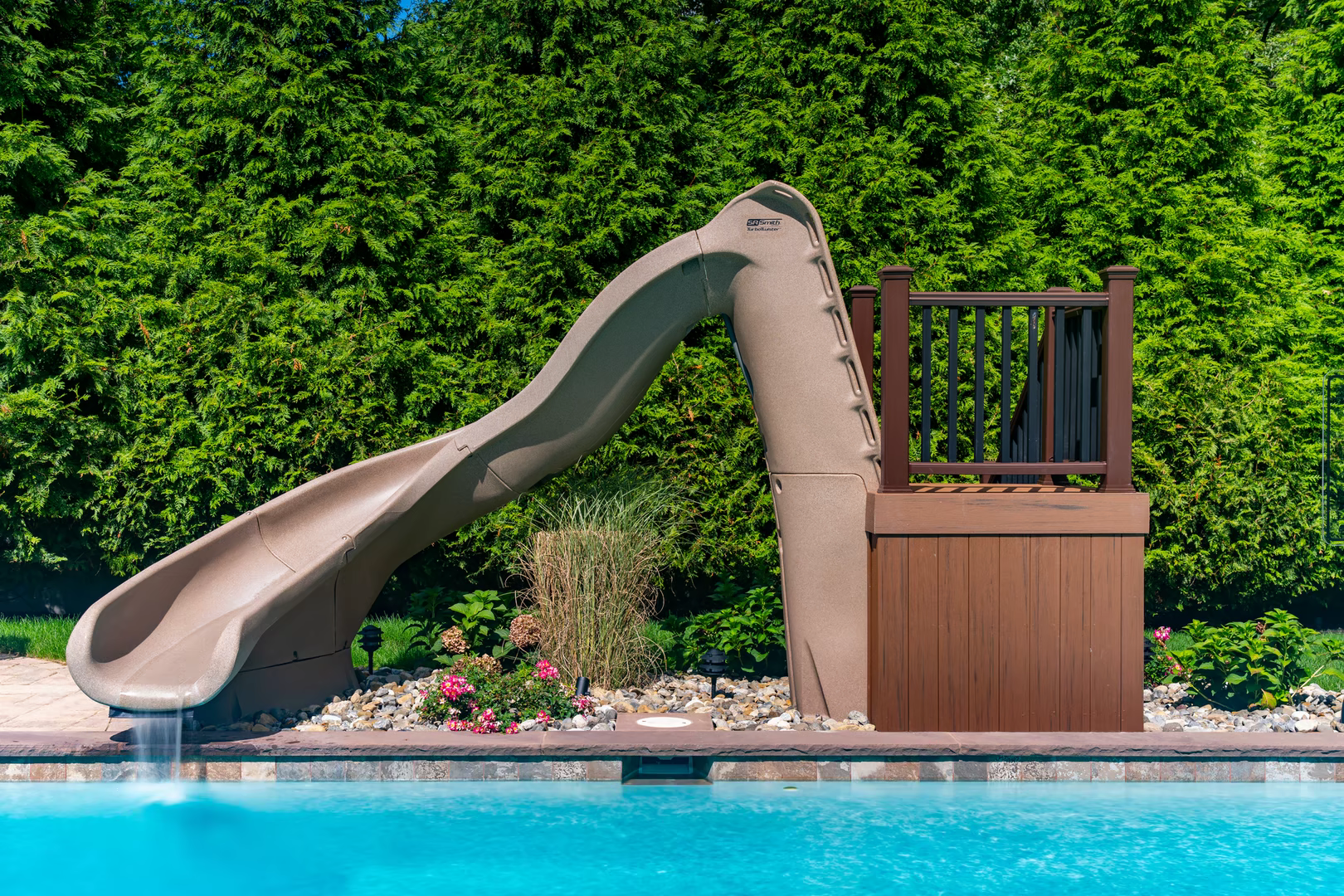 Curved tan pool slide cascading water into a clear pool with lush green landscaping