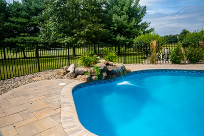 Lemolo C - Hackettstown, NJ - A blue swimming pool with a waterfall in the middle surrounded by greenery and a black fence