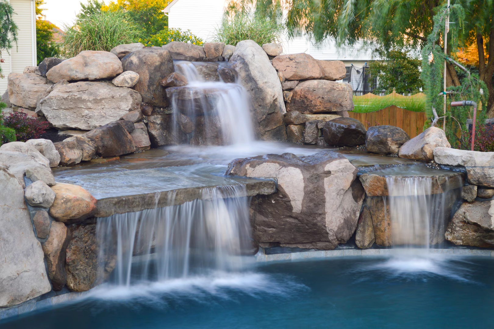 Three-tiered natural stone boulder waterfall cascading into a pool with lush backyard garden plants