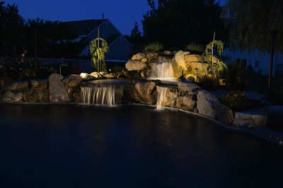 Lisbon M - Marlton, NJ - A pond with a waterfall lit up at night, surrounded by rocks and plants