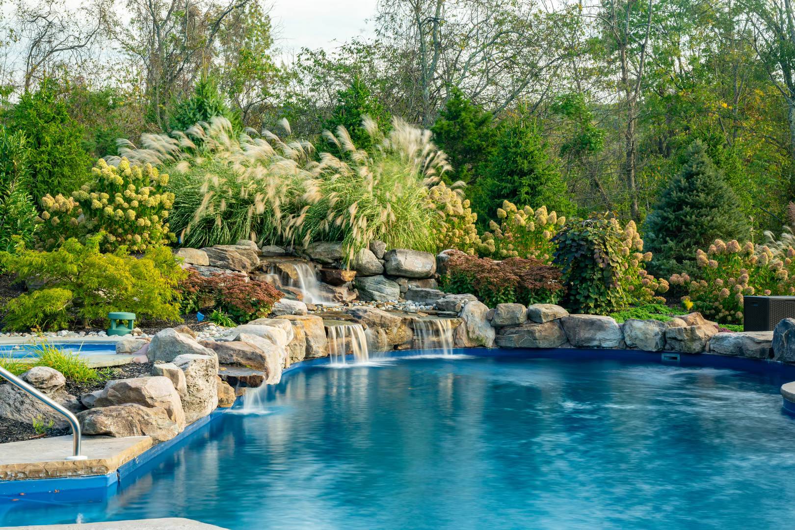Stone boulder waterfalls cascade into a pool surrounded by lush ornamental grasses and plants
