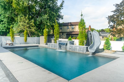Memorial R - Cherry Hill, NJ - A rectangular swimming pool with a waterfall and a stone sculpture in the middle, surrounded by lush greenery and a white fence