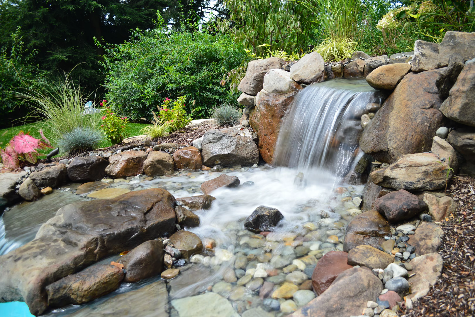 Natural stone boulder waterfall cascading into a pool surrounded by lush garden greenery