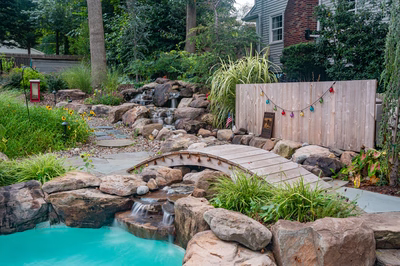 Nachi D - Medford Lakes, NJ - A backyard with a small waterfall and a wooden bridge over a turquoise pool