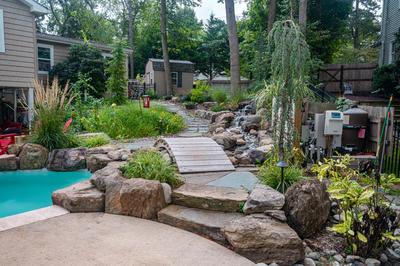 Nachi F - Medford Lakes, NJ - A backyard with a small waterfall and a wooden bridge over a pool surrounded by rocks and plants