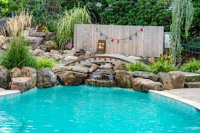 Nachi J - Medford Lakes, NJ - A small wooden bridge over a small waterfall in a swimming pool surrounded by rocks and plants