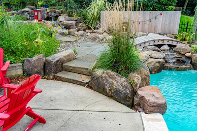 Nachi N - Medford Lakes, NJ - A backyard with a swimming pool surrounded by lush greenery and rocks, with red adirondack chairs and a stone path leading to a small waterfall
