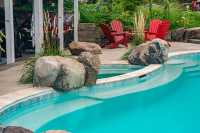 Nachi T - Medford Lakes, NJ - Two red adirondack chairs sit by the edge of a turquoise swimming pool surrounded by rocks and greenery