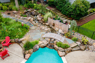 Nachi W - Medford Lakes, NJ - A backyard with a small pool surrounded by lush greenery and rocks, with red adirondack chairs and a wooden bridge