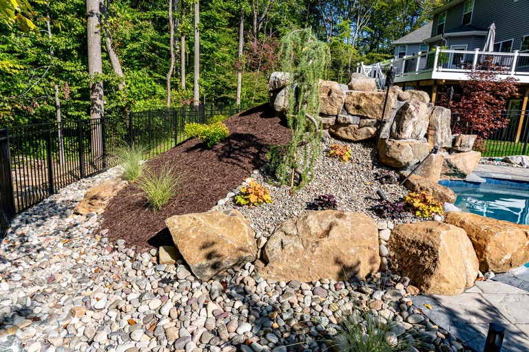 Natural stone boulder waterfall cascading into a pool surrounded by mulch and greenery