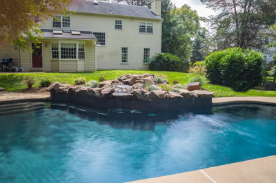 Owen C - Bryn Mawr, PA - A swimming pool with a waterfall in the middle of it surrounded by lush greenery and a house in the background