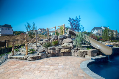 Pearl B - Lincroft, NJ - A backyard with a water feature and a slide, surrounded by lush greenery and houses in the background