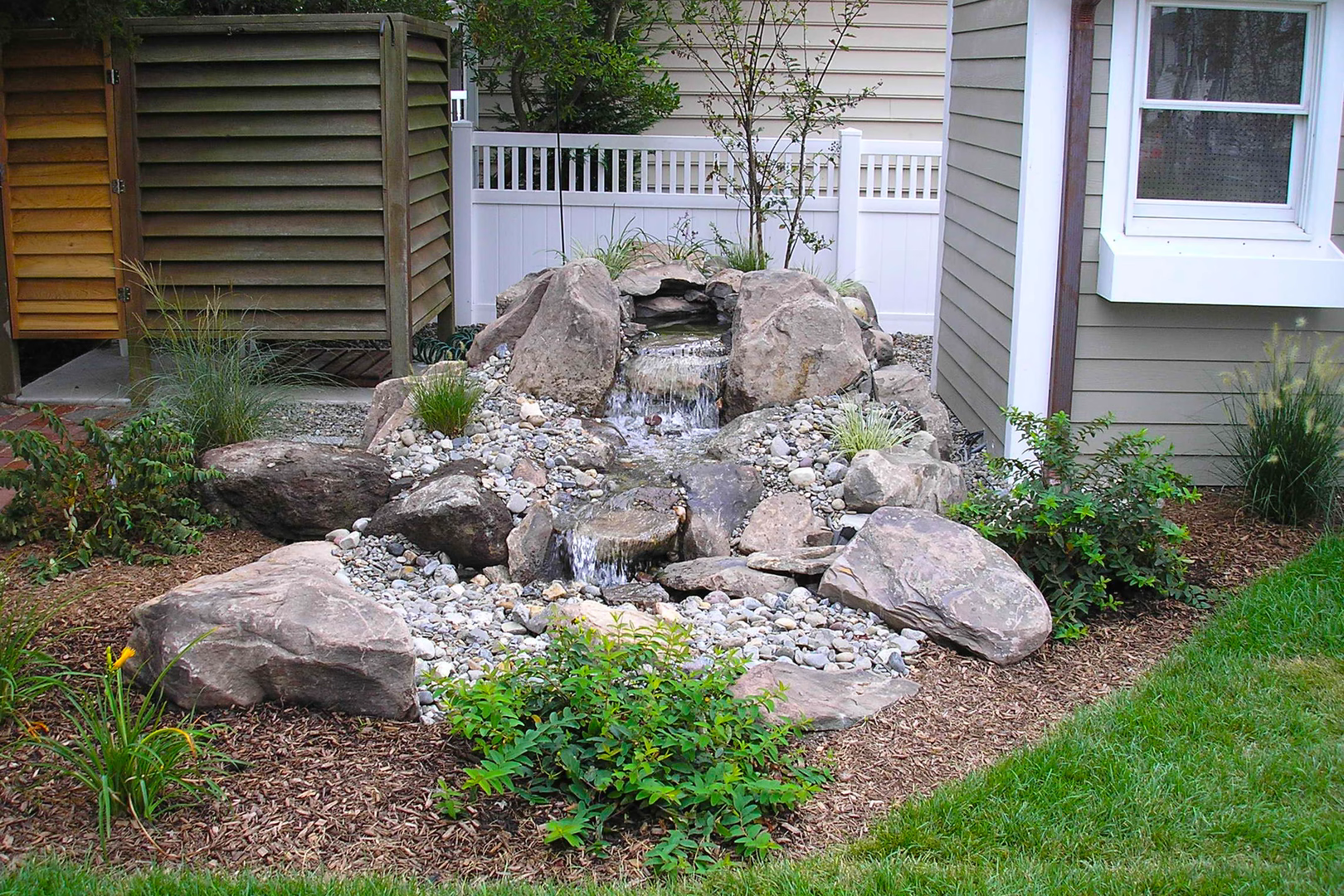 Natural stone boulder waterfall cascading through lush garden plants and decorative mulch beds
