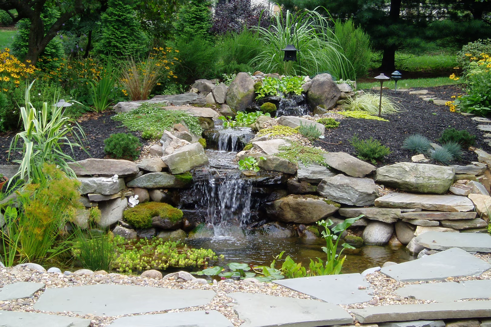 Natural stone boulder waterfall cascading into a pond surrounded by lush garden plants