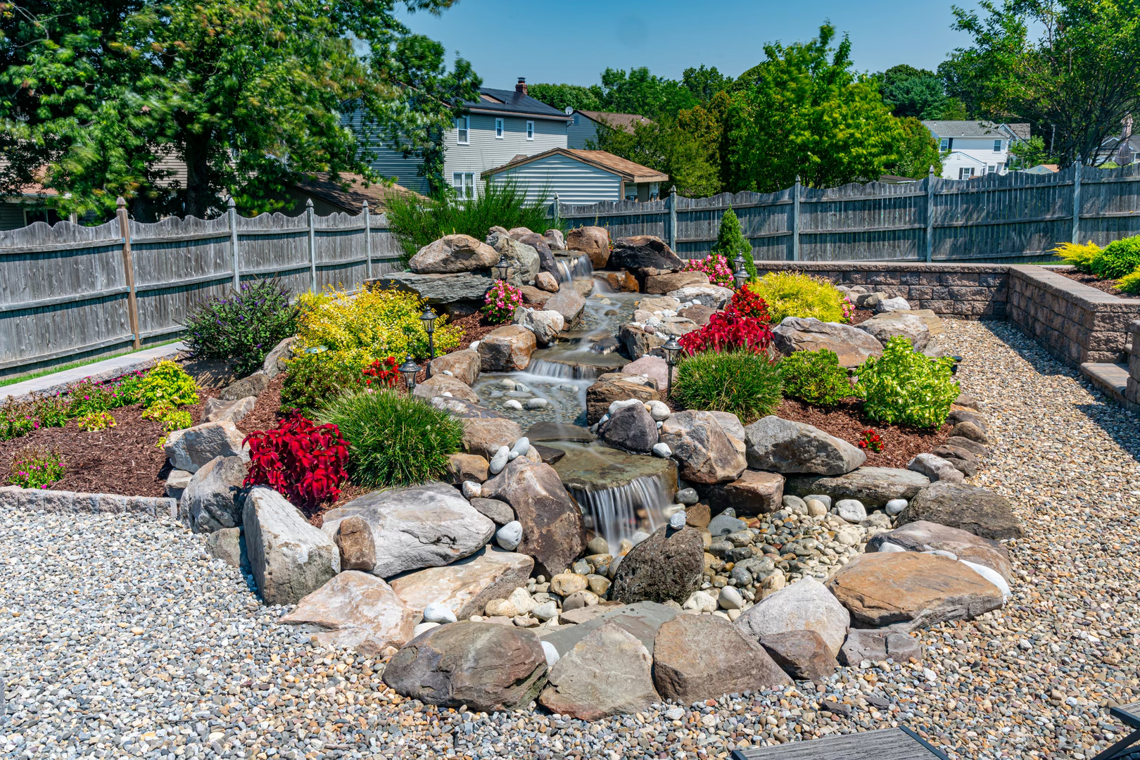 Natural stone boulder waterfall cascading through vibrant plants and decorative gravel landscaping