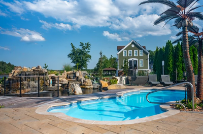Shebelle - Hammonton, NJ - A large swimming pool surrounded by lush greenery and rocks, with a house in the background