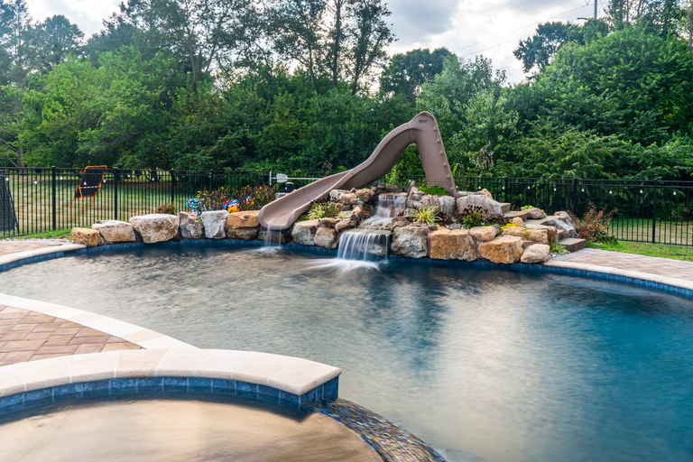 Natural stone boulder waterfall cascading into a pool with a slide and greenery