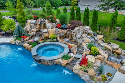 Spray B - Franklinville, NJ - A backyard with a swimming pool and a waterfall feature surrounded by lush greenery and colorful flowers