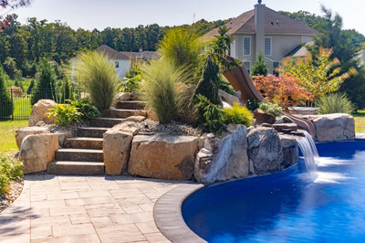 Tamolitch G - Millstone, NJ - A backyard with a swimming pool and a water feature surrounded by plants and rocks