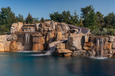 Tchupala F - Medford, NJ - A large rock waterfall with a slide in the middle of a pool surrounded by lush greenery