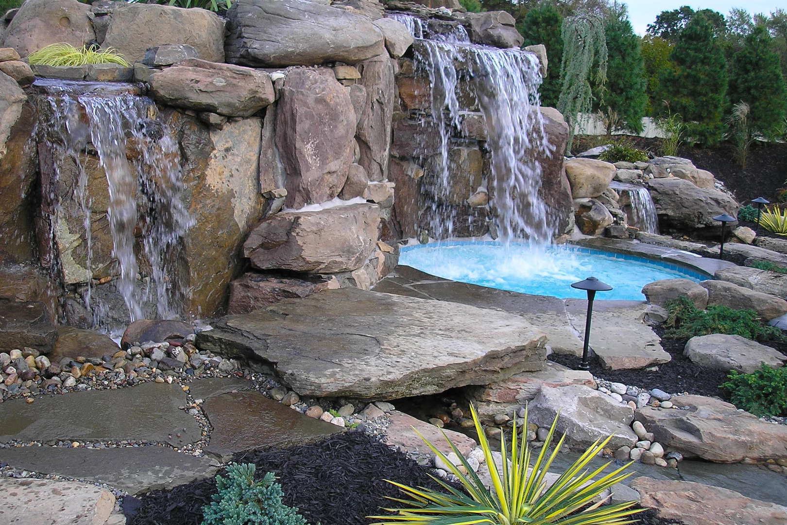 Natural stone boulder waterfalls cascading into a pool surrounded by lush garden plants