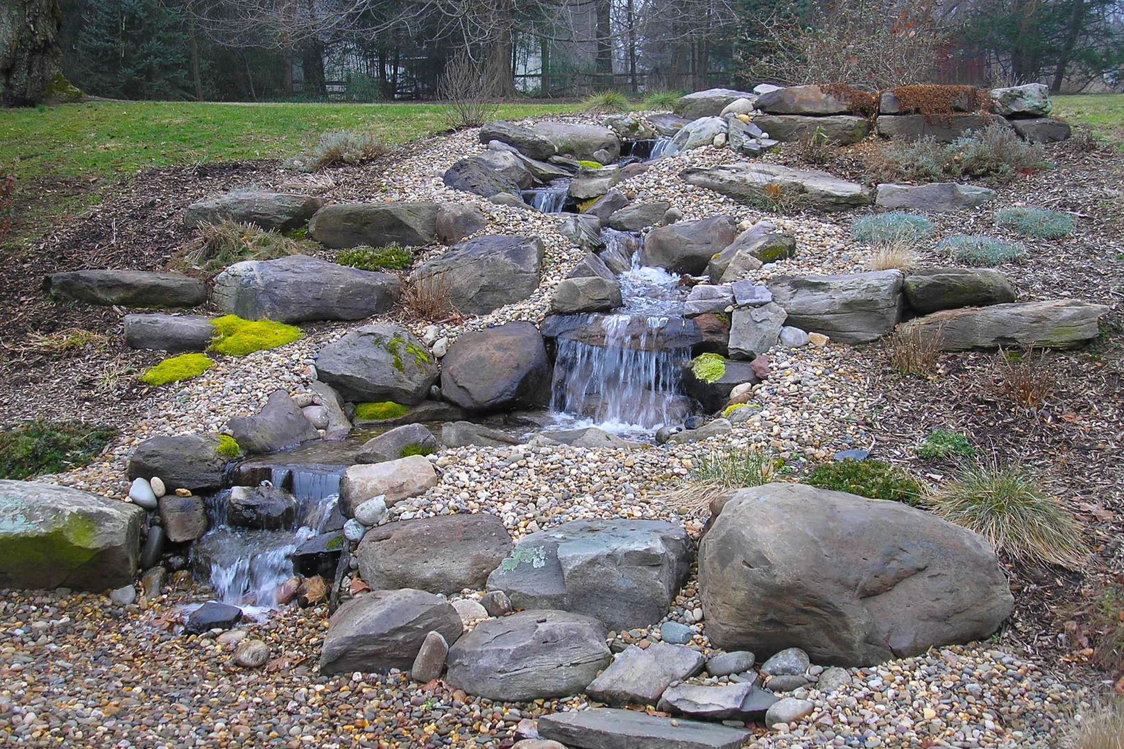 Natural stone boulder waterfall cascading through mossy rocks and lush garden greenery