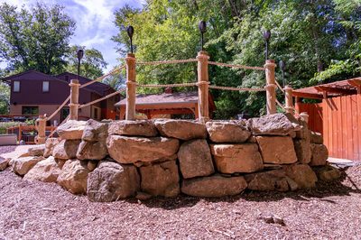 Vernal M - Media, PA - A play structure made of rocks and rope in a wooded area with a house in the background