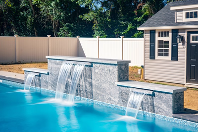 Waireinga A - Bel Air, MD - A swimming pool with a waterfall feature in the middle of it, surrounded by a white fence and a house in the background