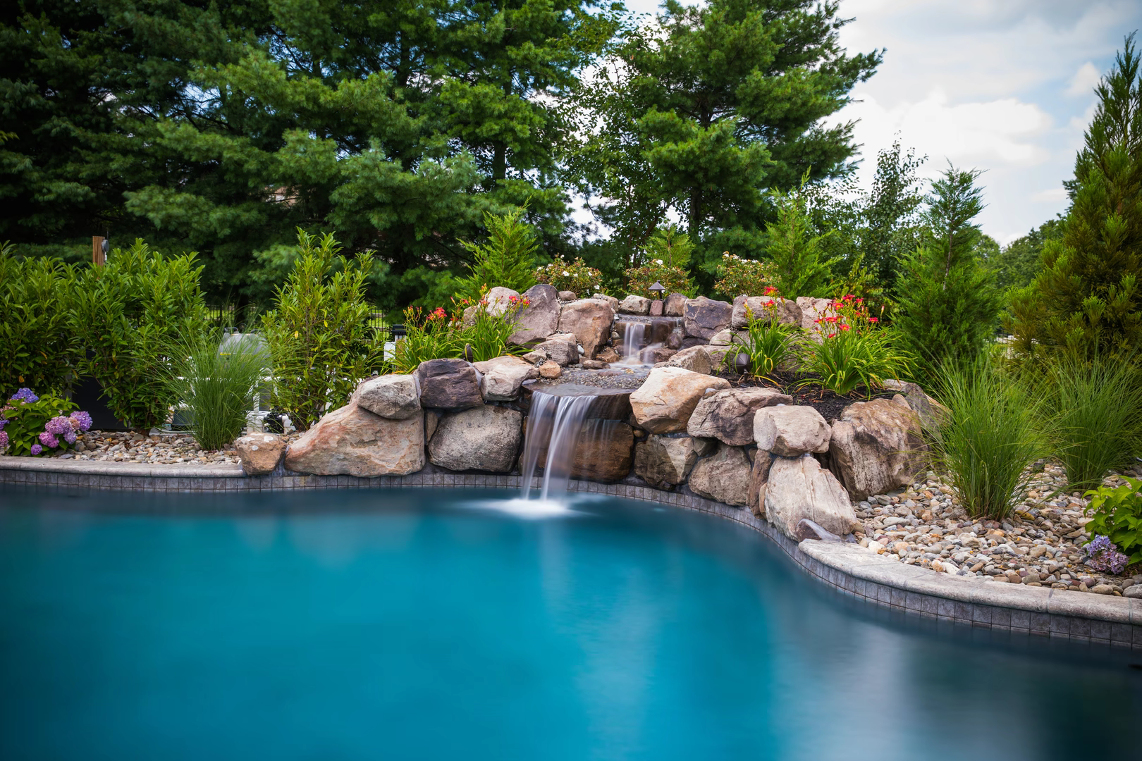 Natural stone boulder waterfall cascading into a pool surrounded by lush garden plants