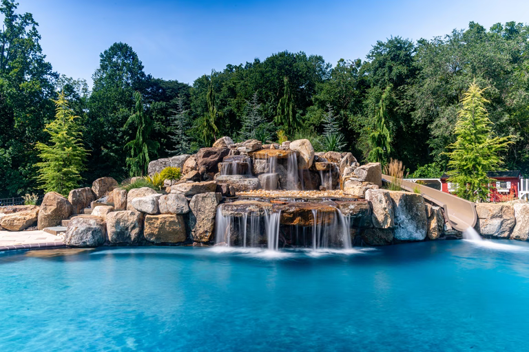 Double-tiered natural stone boulder waterfall cascading into a pool surrounded by lush evergreen trees
