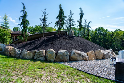 Yakso K - Mullica Hill, NJ - A large pile of mulch on top of a rock wall in a grassy area with trees in the background