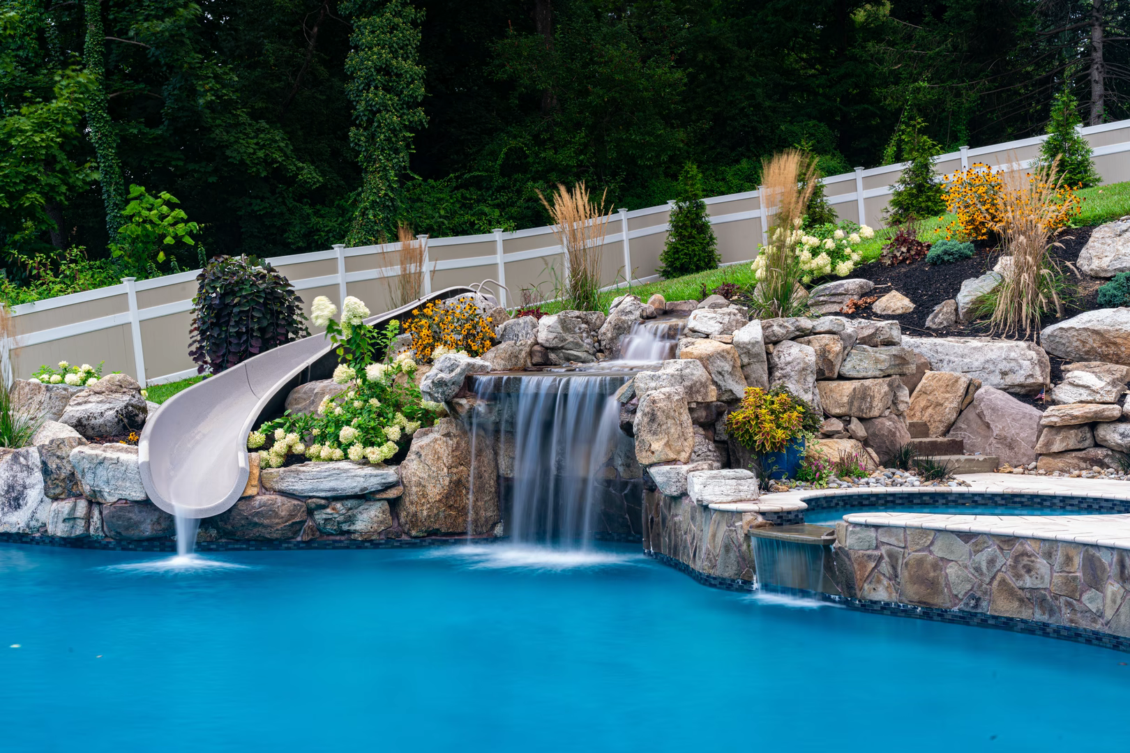 Natural stone boulder waterfall cascading into a pool with lush landscaping and slide