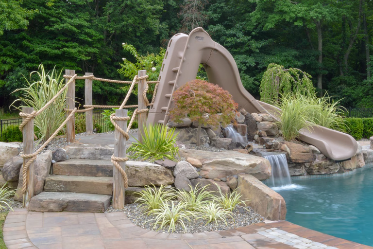 Natural stone boulder waterfall cascading into a swimming pool surrounded by lush greenery