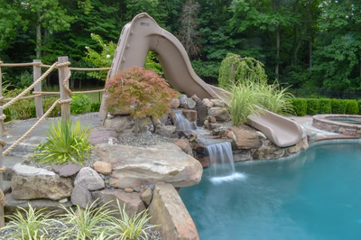 Zillie E - Manalapan, NJ - A backyard pool with a water feature and a slide, surrounded by lush greenery