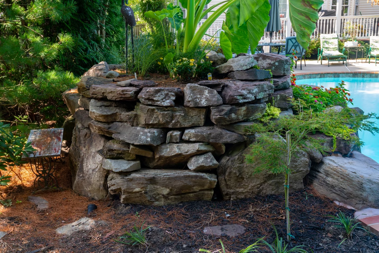 A stone retaining wall next to a swimming pool surrounded by lush greenery
