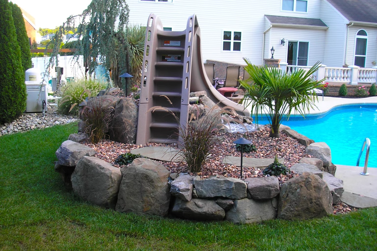 A backyard with a pool and a water slide surrounded by rocks and plants
