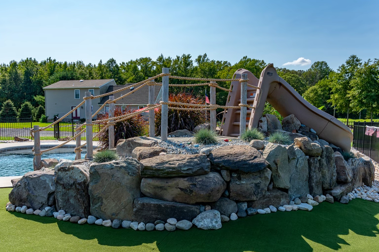 A play area with a rock wall and a slide, surrounded by lush green trees and a house in the background