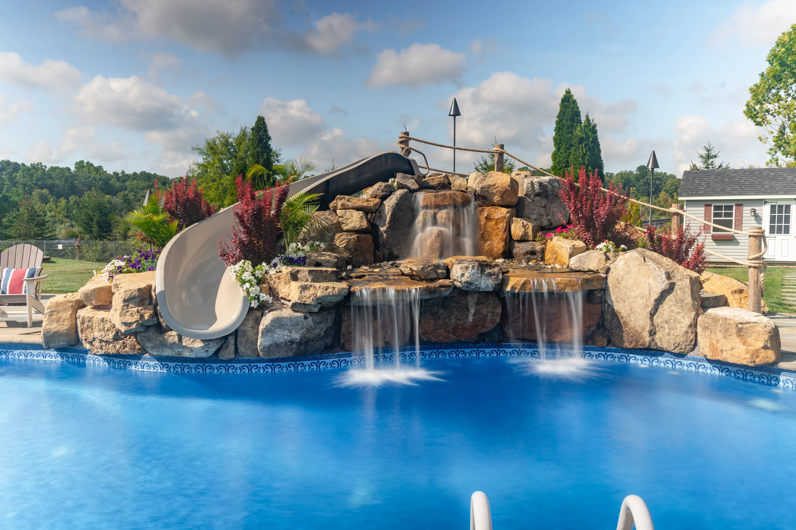 Three-tiered natural stone boulder waterfall cascading into a pool with lush plants and slide