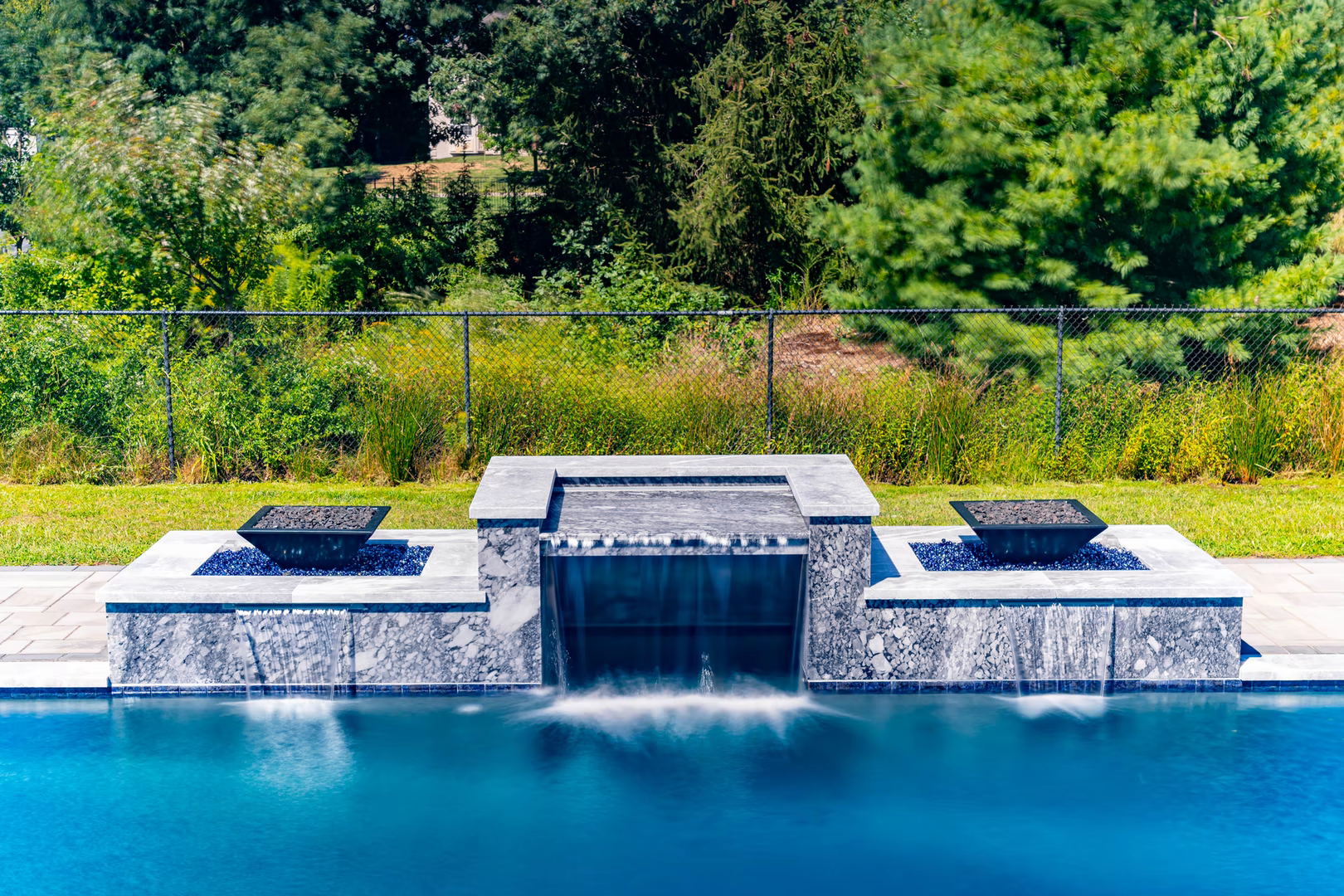 Three-tiered stone waterfall cascading into a rectangular pool surrounded by lush green woodland landscaping