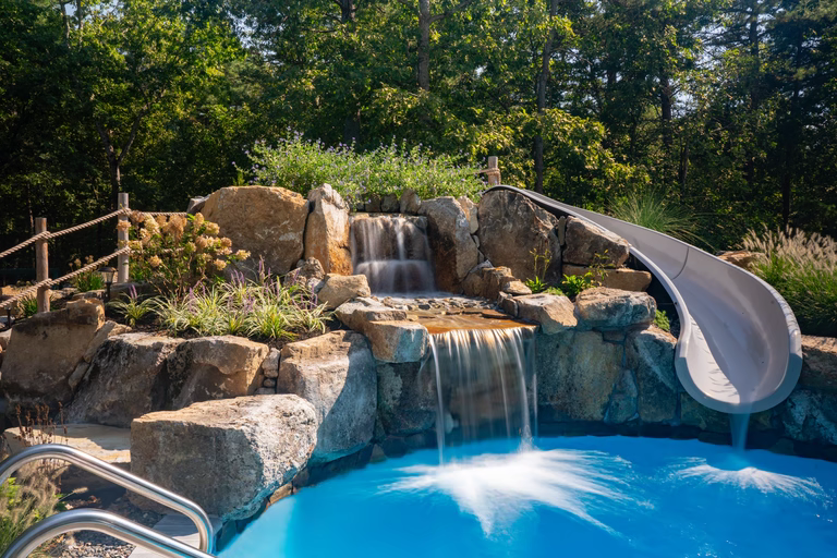 Natural stone boulder waterfall cascading into a pool with lush plants and slide
