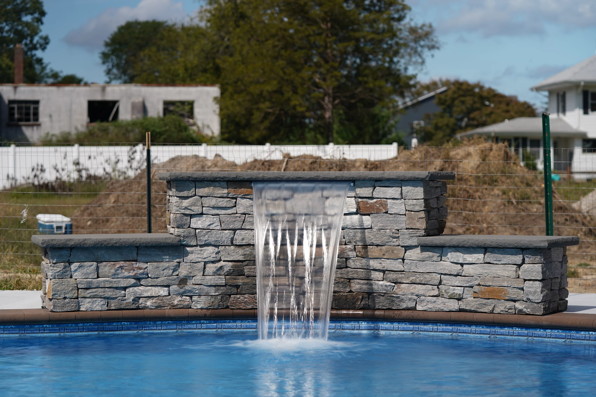 Algonquin A - Hammonton, NJ - This custom installation features a multi-tiered natural stone waterfall cascading into a gunite pool. The waterfall is constructed from irregularly shaped, light gray stone blocks arranged in a tiered design, creating a visually appealing cascade that flows into the pool below. The surrounding area includes a stone bench and a white wire fence, with trees and a partially constructed building visible in the background, indicating a well-maintained outdoor living space.