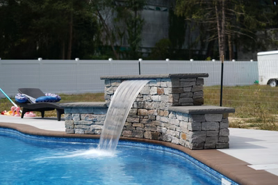 Algonquin D - Hammonton, NJ - This professional installation features a multi-tier natural stone waterfall cascading into a gunite pool. The waterfall structure is constructed from irregularly shaped stone blocks arranged in a tiered design, creating a visually appealing cascade. The surrounding area includes a clean concrete deck, a lounge chair with towels, and a wire fence providing privacy, all set in a well-maintained backyard with trees and a building in the background.