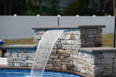 Algonquin E - Hammonton, NJ - This professional installation features a multi-tier natural stone waterfall cascading from a stone ledge into a gunite pool below. The waterfall is constructed with carefully stacked, irregularly shaped stone blocks that create a rustic, organic appearance. The water flows smoothly over the stone surface, creating a soothing visual and auditory effect in the serene backyard setting.