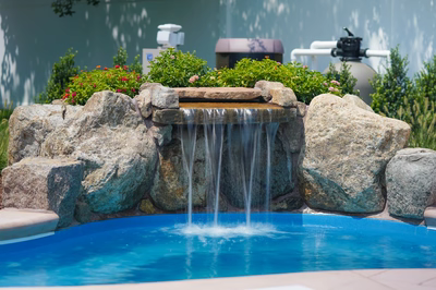 Celilo A - Evesham, NJ - This custom installation features a multi-tier natural stone waterfall cascading into a gunite pool, with large boulders integrated into the design for a natural, rustic look. The waterfall includes a central spillway with multiple streams of water flowing into the pool below, creating a soothing visual and auditory effect. The surrounding area is landscaped with vibrant greenery and flowering plants, enhancing the serene outdoor living space in Evesham, NJ.