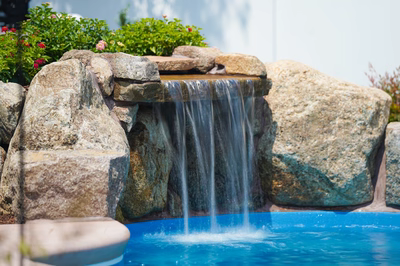 Celilo B - Evesham, NJ - This custom installation features a multi-tier natural stone waterfall cascading into a gunite pool. The waterfall is constructed from large, irregularly shaped boulders arranged in a tiered design, creating a natural, freeform appearance. The pool below has a clean, blue finish and is surrounded by additional natural stone elements, enhancing the rustic aesthetic. The setting includes well-maintained landscaping with vibrant greenery and flowering plants, contributing to a serene backyard environment.